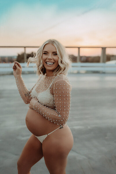 Expecting mother in a parking lot wearing white lingerie and a pearl mesh top.