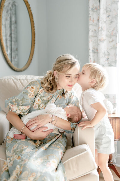 A mother cradles her newborn as her toddler kisses her cheek in their nursery's rocking chair.