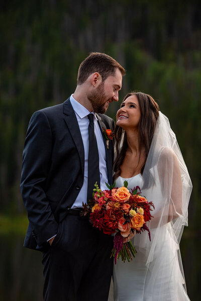 Bride and groom dressed in wedding clothes smiling at each other and holding a fall themed bridal bouquet.