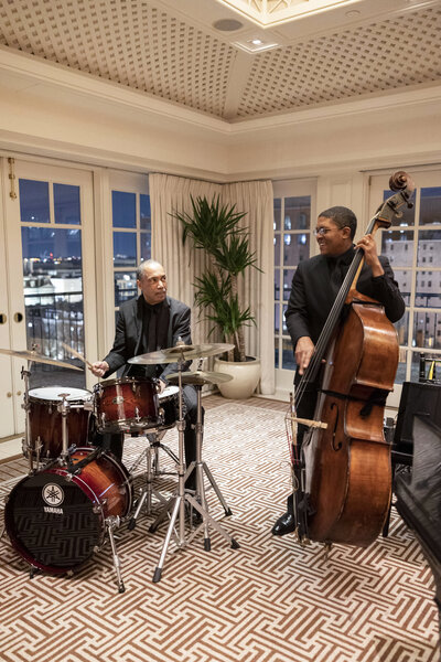 Two musicians perform indoors; one plays the drums and the other plays the double bass. They are in a room with large windows and a patterned carpet. A potted plant is visible in the background.