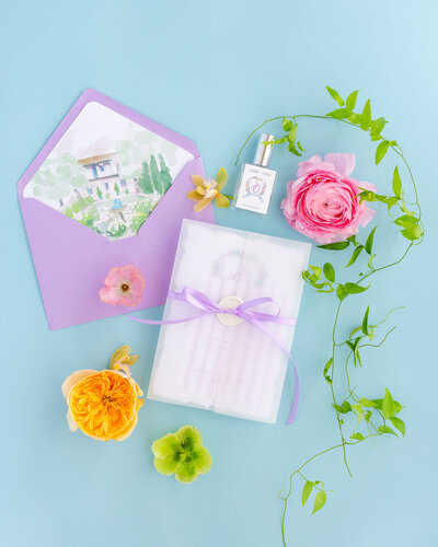 A flat lay of elegant wedding stationery featuring a lavender envelope with a watercolor illustration liner, a wrapped invitation with a purple ribbon, a small perfume bottle, and vibrant spring flowers arranged on a pastel blue background.
