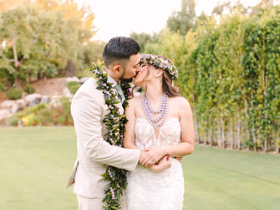 coyote hills golf course bride and groom share a kiss on a grassy lawn. The bride wears a floral crown and lace dress, while the groom is in a light suit with a lei. Romantic and serene.
