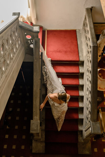 A bride in a lace gown gracefully descending a red-carpeted staircase in a historic chateau, evoking timeless elegance