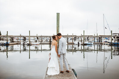 Bride and groom standing on a marina looking back at the camera  at sunset during their Portland Maine elopement with sailboats in the background.