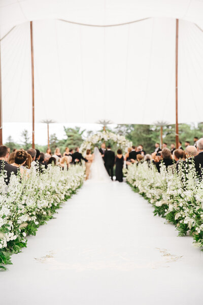 A photo of a wedding ceremony underneath a sailcloth tent, with green and white floral arrangements lining the wide aisle.