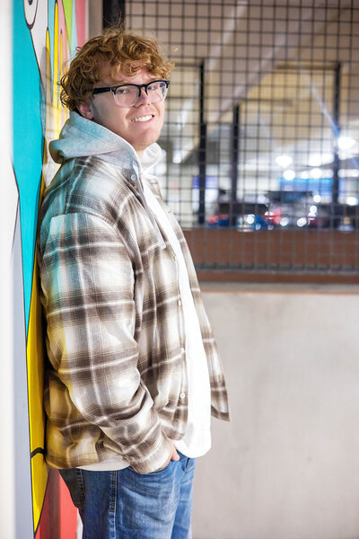 Caucasian senior boy wearing a plaid fleece jacket, white shirt, and blue jeans leaning against a graffiti wall in a parking garage.
