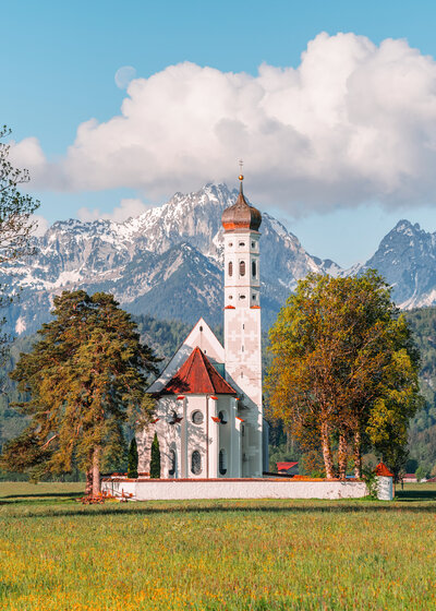 A picturesque white church with a tall steeple surrounded by trees is set against a backdrop of snow-capped mountains under a partly cloudy sky. The scene is peaceful and lush with greenery.