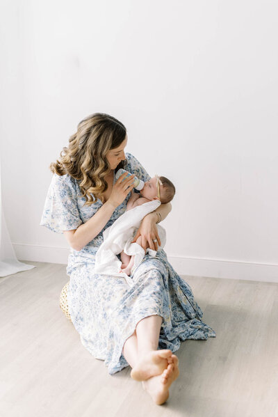  Richmond Newborn Photographer | Photo of a mother in a blue floral dress feeding her newborn baby a bottle in a studio setting 