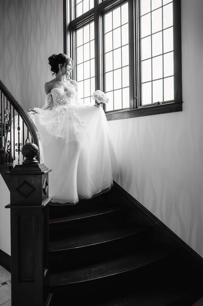 Black and white photo of bride walking down stairs while looking out the window