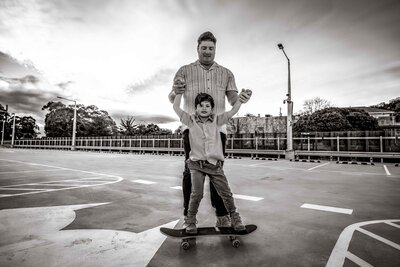 Black and white photo of a little boy standing on a skateboard with his dad behind him holding his hands taken at a rooftop carpark