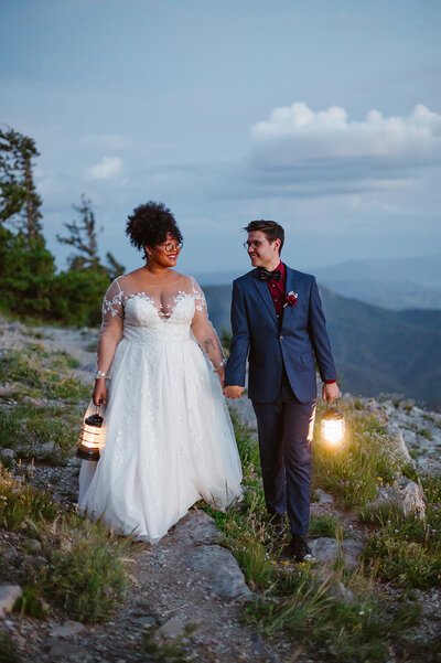 Eloping couple walks hand in hand away from an alpine lake with mountains in the background.