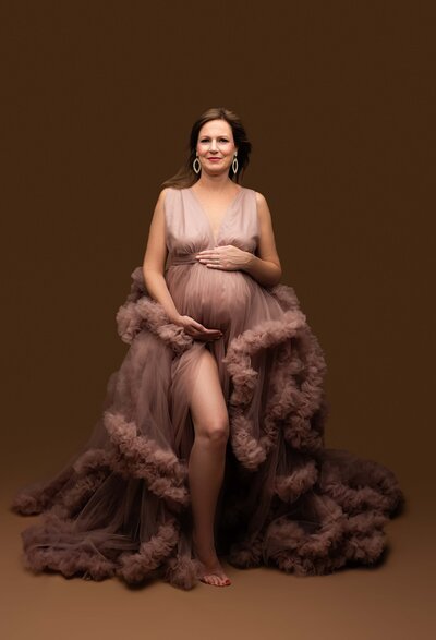 Pregnant women in pink tulle gown on brown backdrop looking into the photographers camera with hair blowing in wind