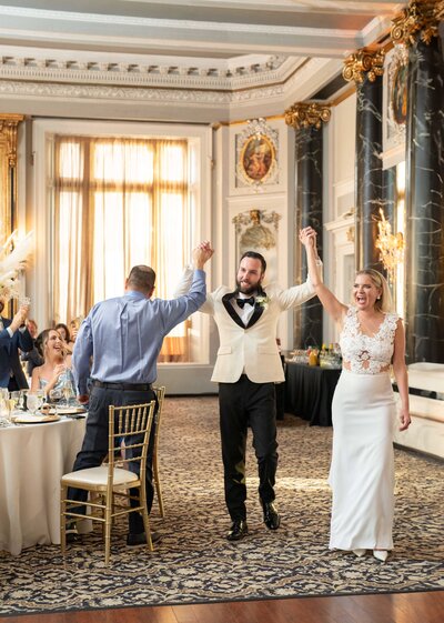A joyous couple in wedding attire, holding hands and celebrating with a guest in an ornate ballroom. The room features tall windows, decorative pillars, and a table with seated guests on the side.