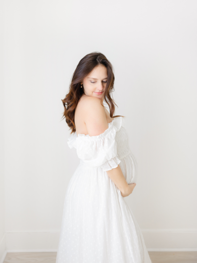pregnant woman wearing simple white dress holds belly and looks out the window