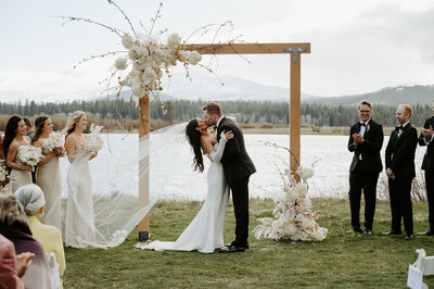 The newlyweds share a kiss beneath their arbor, with a stunning mountain backdrop framing the moment in natural grandeur.