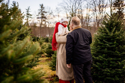 Little baby dresses in a white bear snowsuit and red scarf being lifted and cooed at by mom and dad at a christmas tree farm session