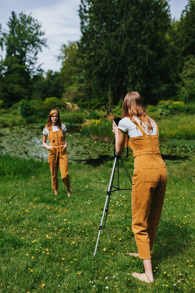 Photographer stands behind camera on top of tripod wearing a mustard-colored jumpsuit taking a photo of herself posing in front of the camera outdoors.