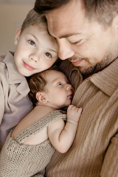 Father holding newborn baby close to his chest while big brother gently leans in during an in-home family photography session in Las Vegas by Angela Wolf Photography