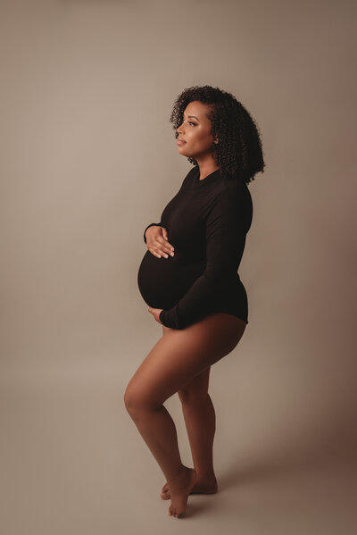 3rd trimester pregnant woman posing in a Woodstock GA maternity portrait studio wearing a black long sleeve body suit  of her while she shows her baby bump. Shot on gray backdrop