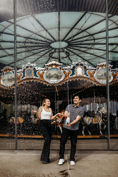Engaged couple opens a bottle of champagne and sprays it in front of the Brooklyn Bridge Park carousel