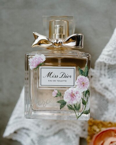 A square bottle of Miss Dior perfume .  It has a cap with a gold bow.  It is laying flat on a ribbon of  white lace.  The bottle has  a small hand painted peony in the top left corner, and 2 bigger peonies and a bud with green leaves and stems in the bottom corner of the bottle.  It  adds an extra dose of luxury to  this lovely gift.