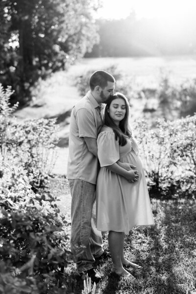 Husband and wife hug and hold baby bump during summer maternity session in Hollis, new Hampshire