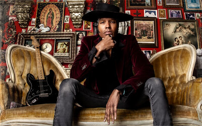 Musician portrait Jeau James sitting on gold sofa guitar beside him resting chin on fist wearing black hat burgundy blazer jeans  religious photos on wall behind him