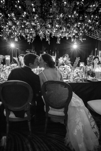Two newlyweds kissing at a decorated table beneath hanging lanterns and foliage at their wedding reception at Hotel del Coronado.  This wedding was planned and designed by Thater Made, a luxury wedding and event producer based in San Diego, CA.