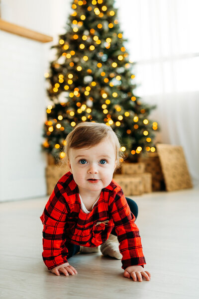 Young child in red plaid sitting beside a decorated Christmas tree during Ottawa holiday mini session.