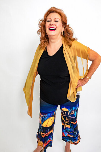 A woman with reddish hair laughs joyfully against a white background. She wears a black top with a sheer gold wrap or scarf, and colorful patterned pants featuring blue and yellow artistic designs.
