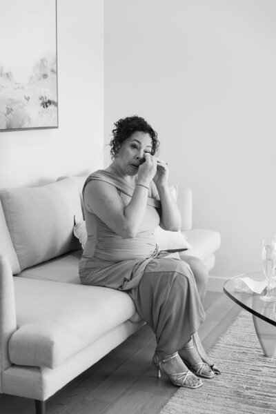 The Lakehouse Inn MA  | Kelsey Sheehan Photography Timeless Rhode Island Weddings | A woman in a formal dress sits on a sofa, applying makeup in a calm and focused manner. The room is elegantly furnished, conveying a serene atmosphere.