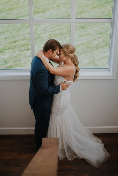 A couple embraces intimately by a large window in a Leesburg VA wedding venue. The person on the right wears a white, strapless gown, and the person on the left is in a dark suit. Outside, a lush green field is visible. The setting is bright with natural light.
