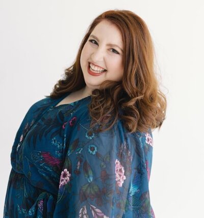 A woman with medium-length red hair smiles widely at the camera. She is wearing a blue dress with a floral pattern, and the background is a plain white, giving the image a clean and bright appearance.