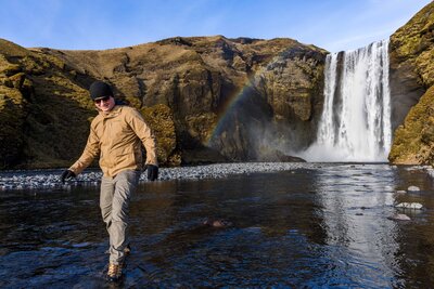 Two grooms stand on a flat rock in front of a large cascading waterfall. The waterfall is falling into a natural pool behind them. They are standing facing each other while reading vows and looking lovingly at one another. 