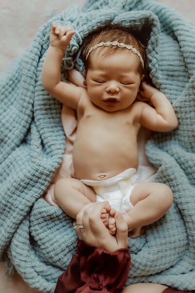 A newborn baby wearing a headband and diaper sleeps, swaddled in a blue blanket with their arms up and legs crossed. Perfect for capturing timeless baby photos in Macon, GA.
