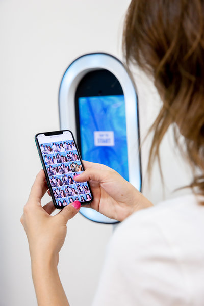 a womans hands holding a phone with photos on the screen in front of a white photo booth