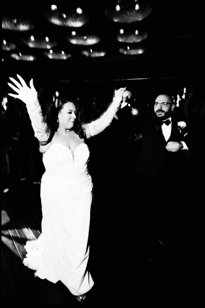 A bride in a white gown raises her arms joyfully while holding hands with a smiling groom in a suit as they dance together under glowing lights at their wedding reception, beautifully captured by a talented NJ wedding photographer.