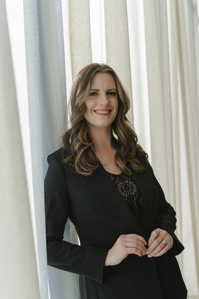 Southern California wedding planner in a black suit standing against a window with a white curtain.