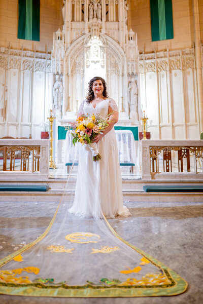 Bride standing in a church with a colorful bouquet and a long embroidered veil