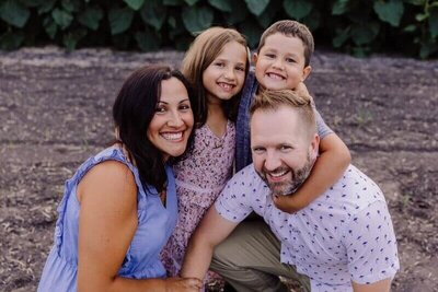 A joyful family of four crouches down together, all beaming with big smiles in front of green foliage.