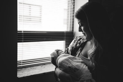 new mother holding newborn bab y near hospital window with black and white conversion