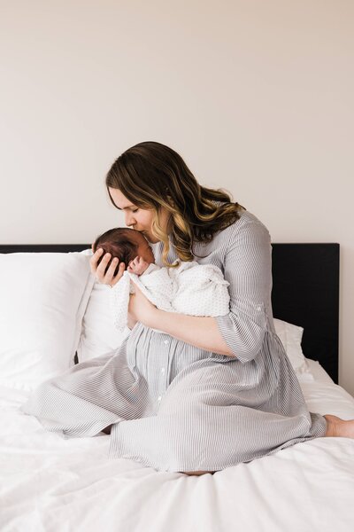 A woman gently cradles and kisses a newborn baby during an at-home newborn photography session while sitting on a bed.
