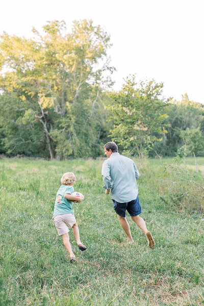 Young boy runs holding football with dad in green field