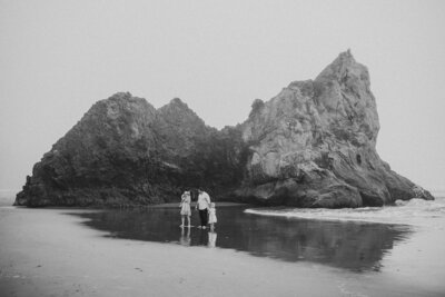 Black and white photo of a family standing barefoot on the beach in front of large coastal rock formations, sharing a quiet moment together.