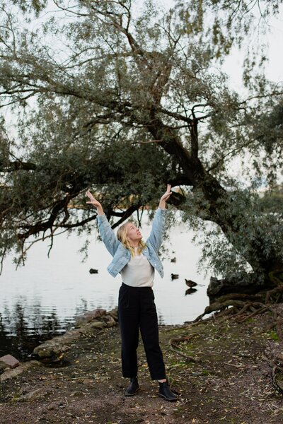 mielen paremminvoinnin ja kehomieliyhteyden valmentaja  seisoo järven rannalla puiden keskellä käden kohotettuina ilmaan