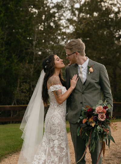 A bride in a lace gown and veil lovingly gazes at the groom in a gray suit holding a vibrant bouquet. They stand on a path with trees in the background.
