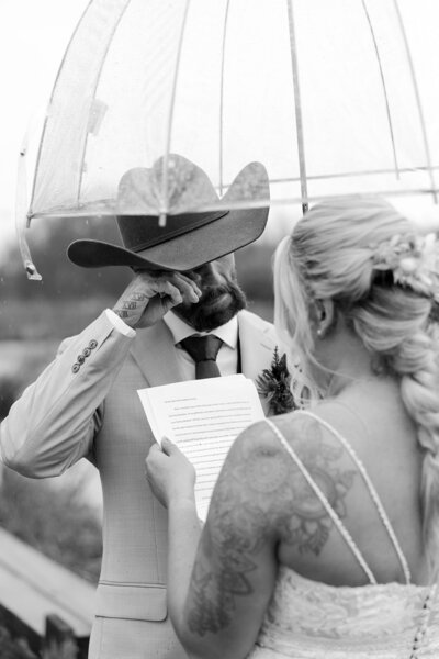 Black and white photo of a bride reading her vows to a crying groom under an umbrella.