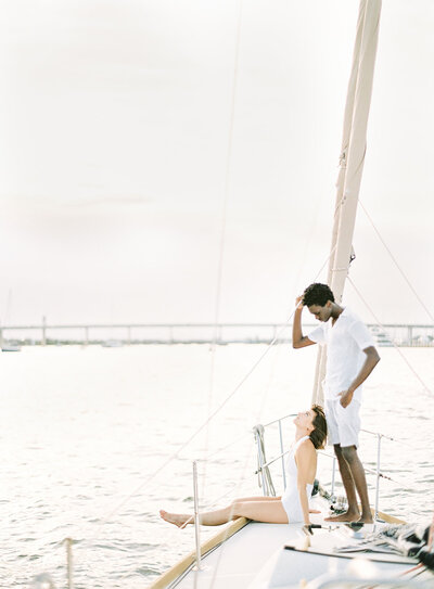 Engagement session on a sailboat. She is wearing a white halter top swimming suit sitting on the side of the boat with one leg down towards the water and the other one lifted straight out as she is kicking her legs and looking up at him. He is wearing white shorts with a white short sleeved dress shirt, standing behind her looking down at her while holding on the the rope of the sail. You can see water and a bridge behind them. Photographed by wedding photographers in Charleston  Amy Mulder Photography