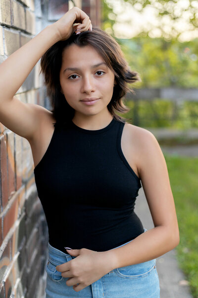 Senior girl with black hair in black tank top and blue jeans leaning against graffiti wall at sunset