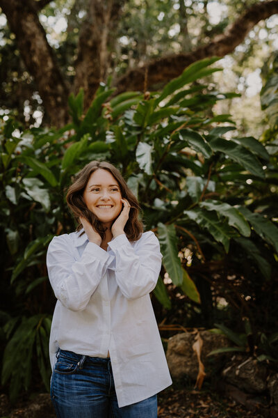 Brandi of the Intuitive Momma wears jeans and a white button down. She smiles and holds her hands around her face with big, leafy plants out of focus behind her.